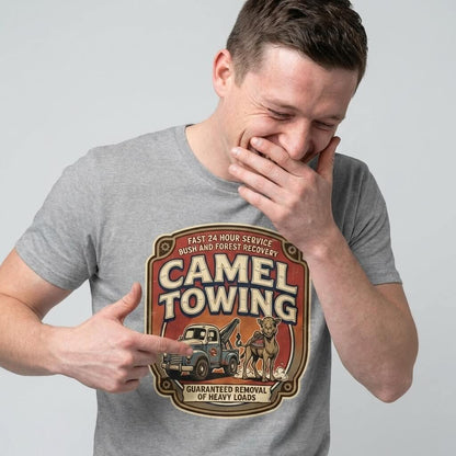 A man in a gray t-shirt laughs while pointing at his Camel Towing Funny Tee, which features a camel, a tow truck, and the phrase "Guaranteed removal of heavy loads.
