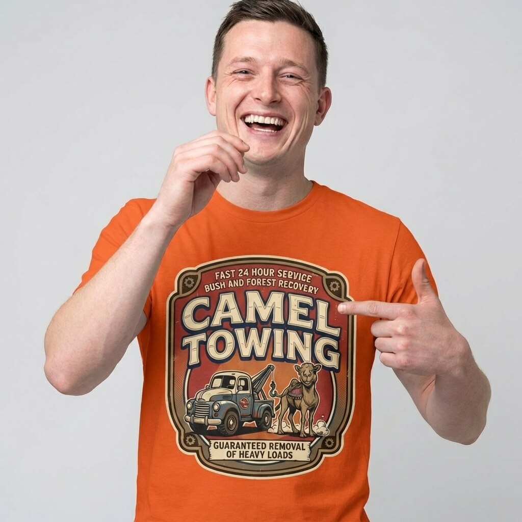A smiling man wears the Camel Towing Funny Tee, featuring a tow truck, camel, and "Guaranteed removal of heavy loads" text. He points at the graphic and laughs—making this novelty tee a perfect gift for anyone with a sense of humor.