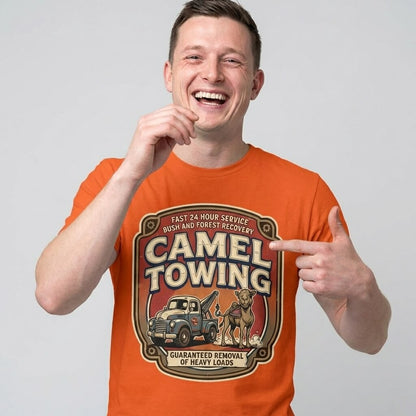 A smiling man wears the Camel Towing Funny Tee, featuring a tow truck, camel, and "Guaranteed removal of heavy loads" text. He points at the graphic and laughs—making this novelty tee a perfect gift for anyone with a sense of humor.