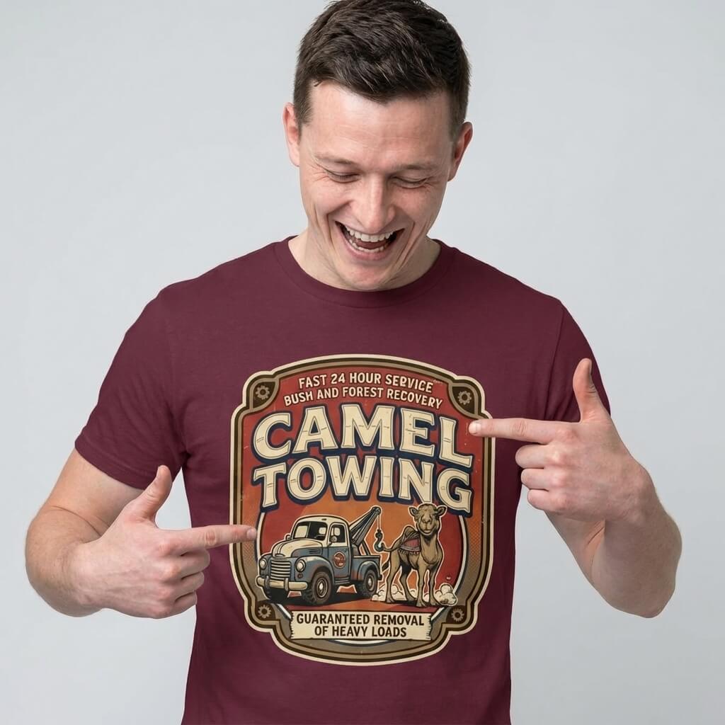 A smiling man points at his maroon Camel Towing Funny Tee, featuring a cartoon tow truck and camel with “Camel Towing: Guaranteed Removal of Heavy Loads”—the perfect novelty graphic t-shirt gift for anyone who loves humor.