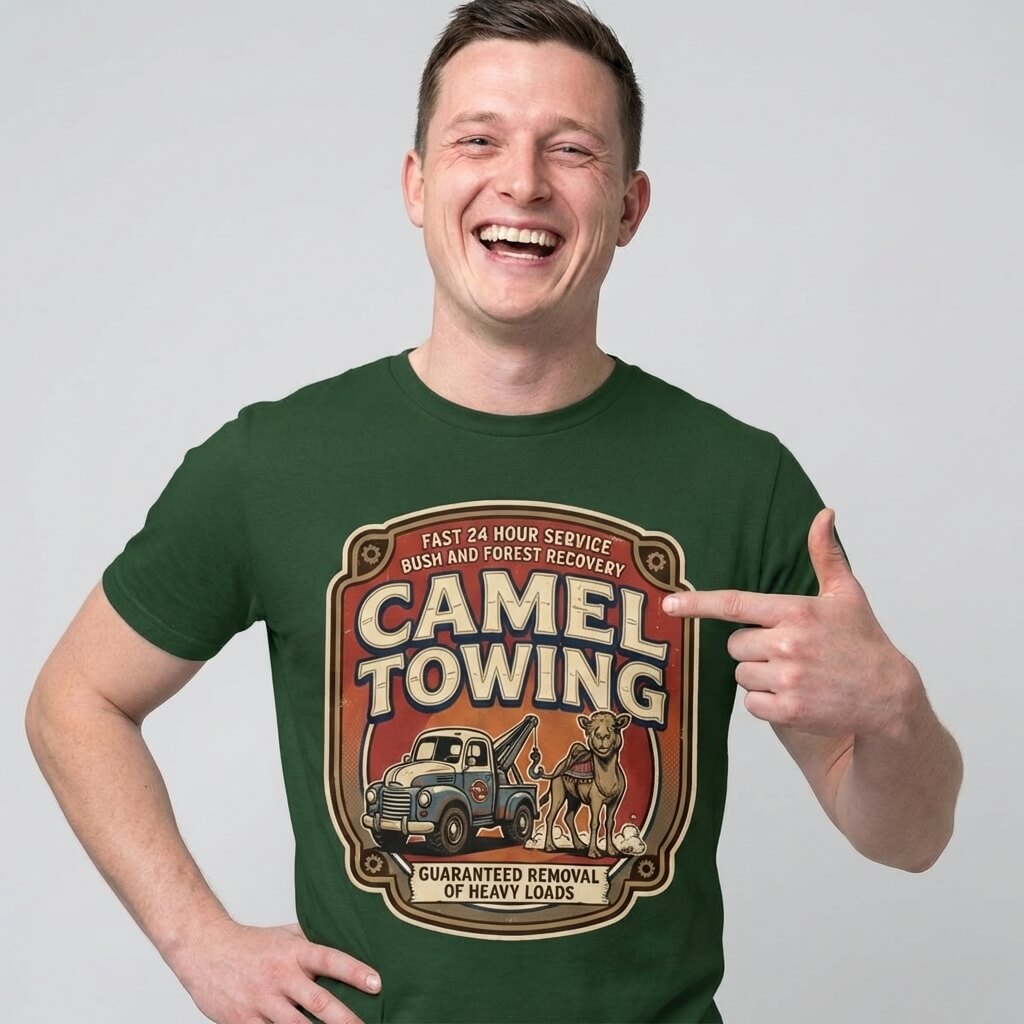A smiling man in a green Camel Towing Funny Tee points to its tow truck and camel graphic with the text “Camel Towing: Guaranteed Removal of Heavy Loads”—the perfect novelty gift for fans of this hilarious shirt.