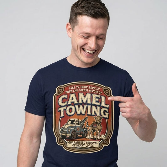A smiling man shows off his Camel Towing Funny Tee—a novelty graphic t-shirt with a camel and tow truck design and the phrase: Guaranteed Removal of Heavy Loads, Fast 24 Hour Service—great as a gag gift.