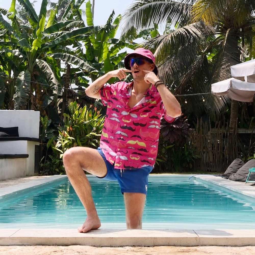 Wearing blue shorts, a bright pink mustache-patterned shirt, and the Movember Bucket Hat, a man poses playfully by a swimming pool surrounded by lush tropical plants and palm trees.