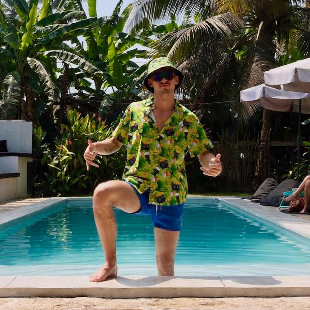 Wearing a St Patrick’s Day Bucket Hat, a man in a colorful shirt and blue shorts stands by the poolside giving thumbs up, surrounded by lush tropical plants and palm trees.