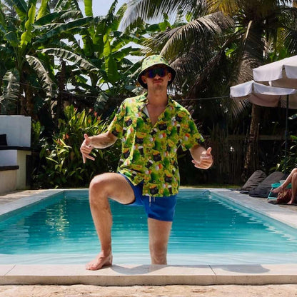 Wearing a St Patrick’s Day Bucket Hat, a man in a colorful shirt and blue shorts stands by the poolside giving thumbs up, surrounded by lush tropical plants and palm trees.