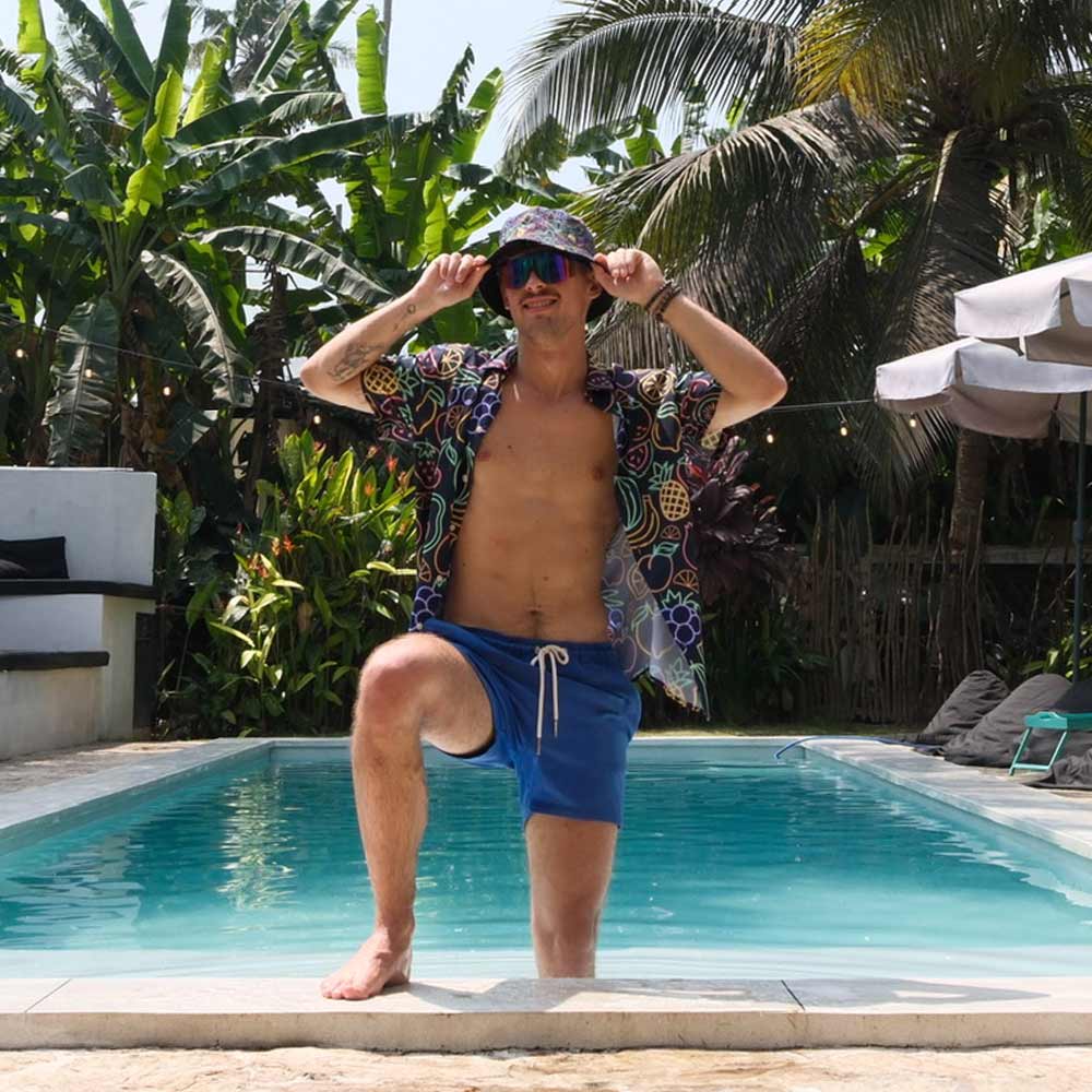 A man wearing a colorful shirt, blue shorts, and the Fluorescent Fruit Fiesta Bucket Hat poses by a swimming pool edged with lush green tropical plants and palm trees.