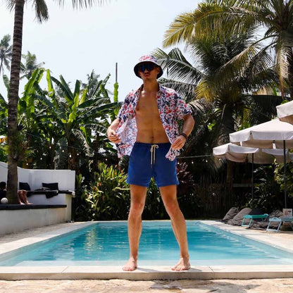 A man stands confidently by a pool surrounded by tropical plants, wearing sunglasses, blue swim shorts, an open floral shirt, and the England Football Bucket Hat.