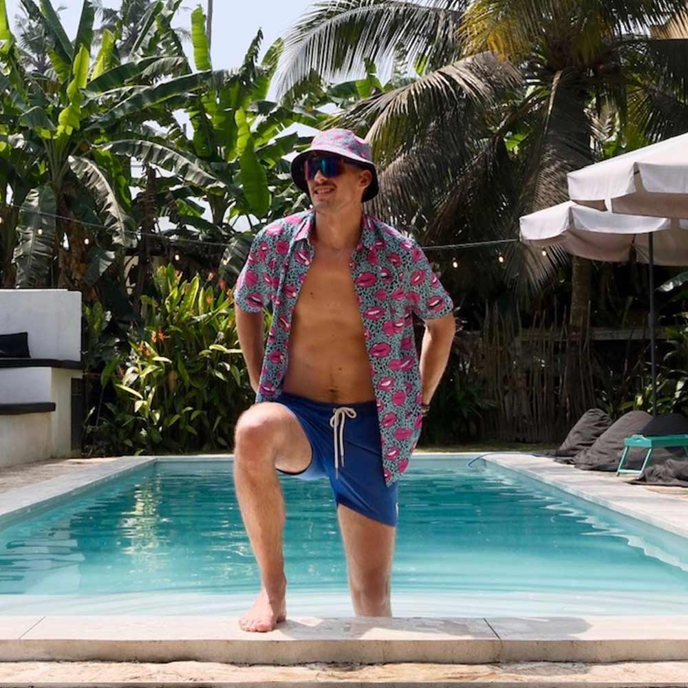 A man in a colorful shirt, blue shorts, and the Leopard Kiss Bad Bucket Hat stands by a pool, smiling with tropical plants and palm trees behind him.