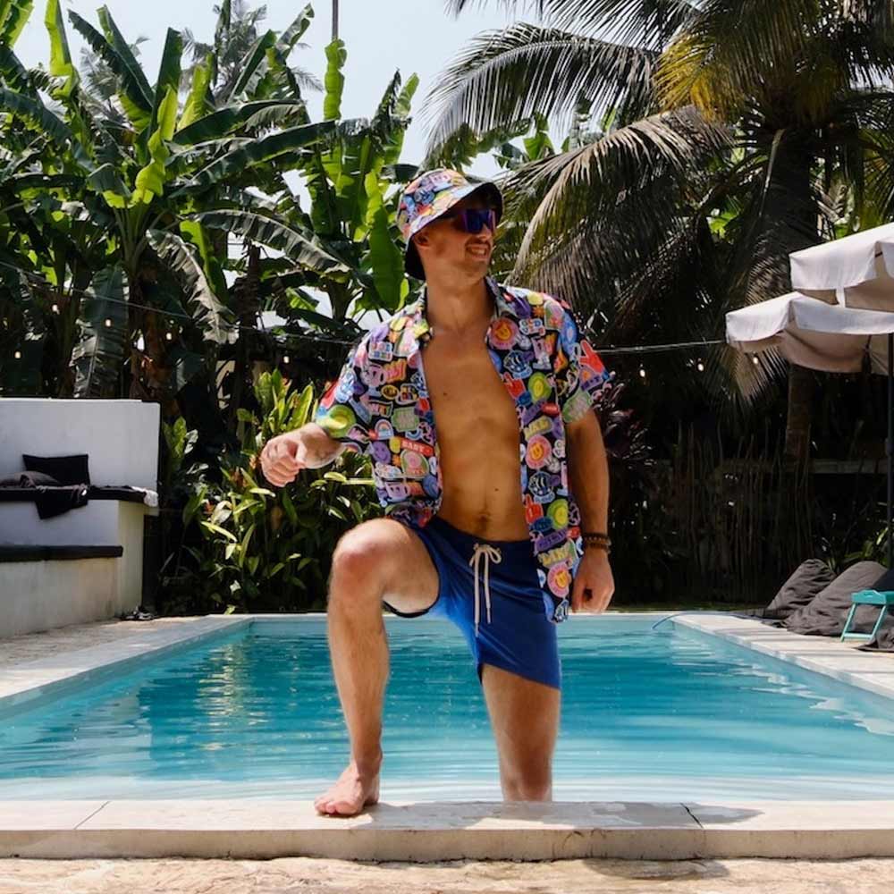 A man in a colorful shirt, blue shorts, and the Retro Sticker Bucket Hat stands poolside with one knee raised. He smiles in sunglasses, surrounded by tropical plants and palm trees in the background.