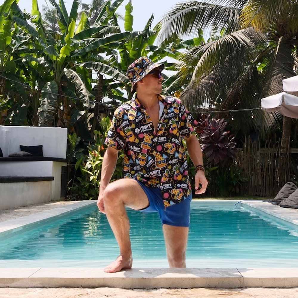A man in a colorful Sh!t Shirt Club shirt, the Wanna Pizza This Bucket Hat, and blue swim trunks poses with one knee on the pool edge amid tropical plants and palm trees on a sunny day.