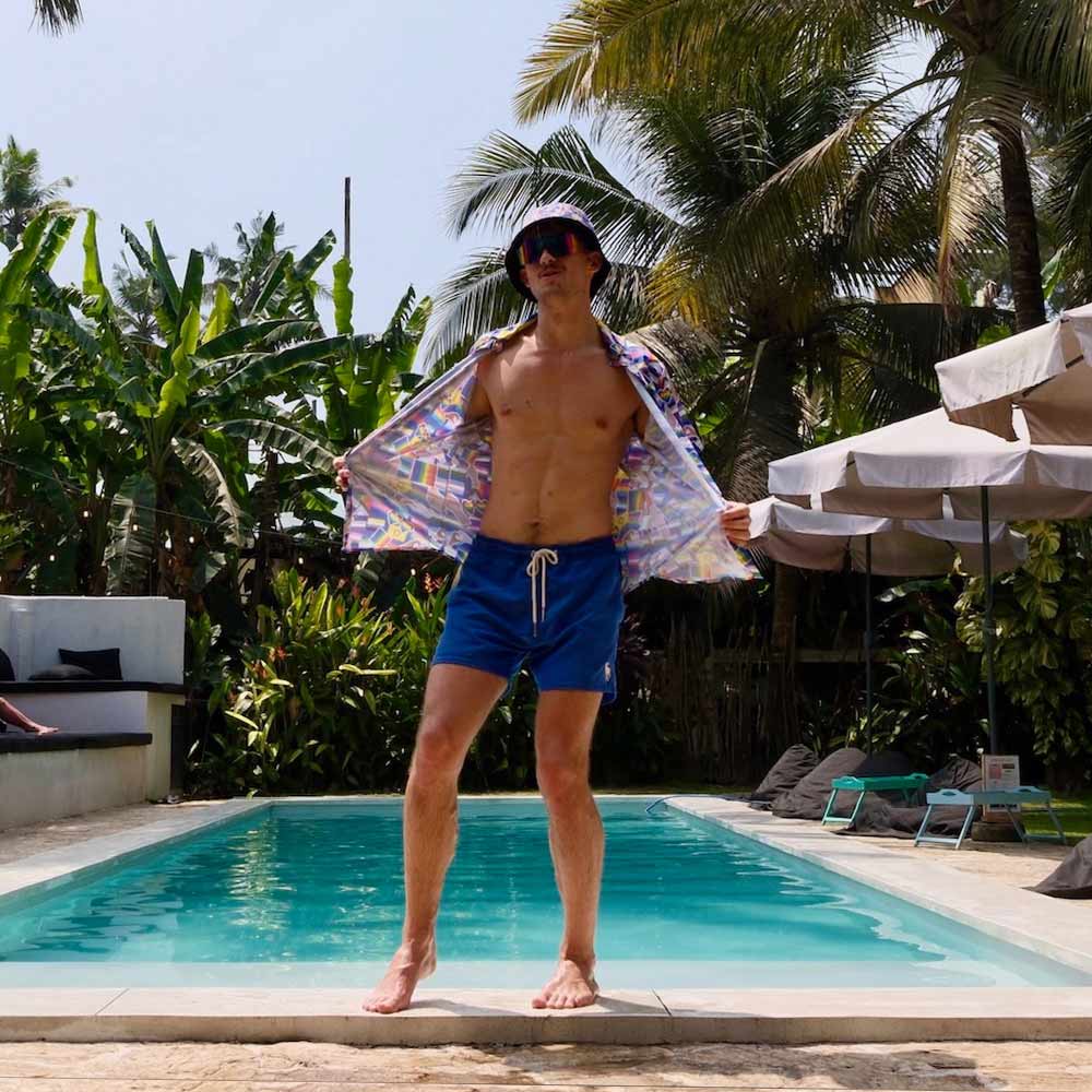 A man in blue swim shorts, sunglasses, and an open patterned shirt confidently poses by a pool under the sun, wearing the LGBTQ Pride Bucket Hat amid tropical plants and palm trees.