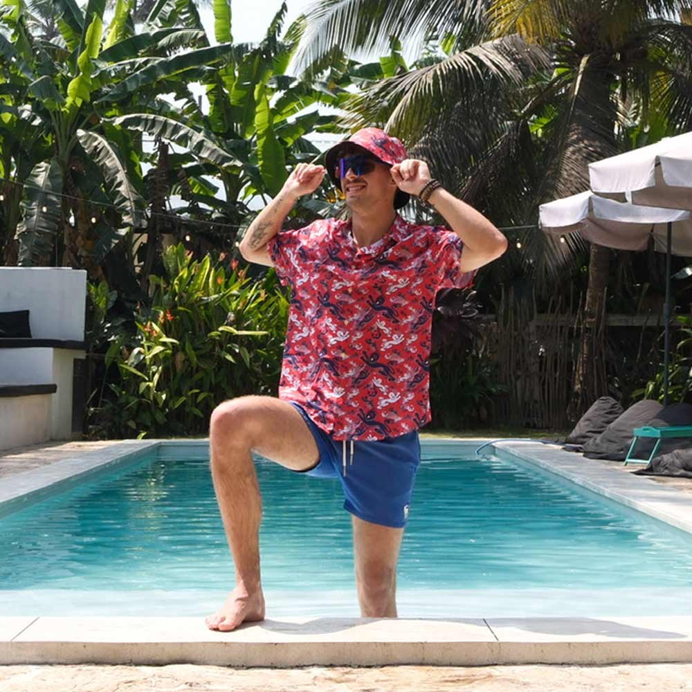 Wearing the Red England FC Hawaiian Shirt, sunglasses, blue shorts, and a matching hat, a person poses playfully by a pool amidst lush tropical plants and palm trees.