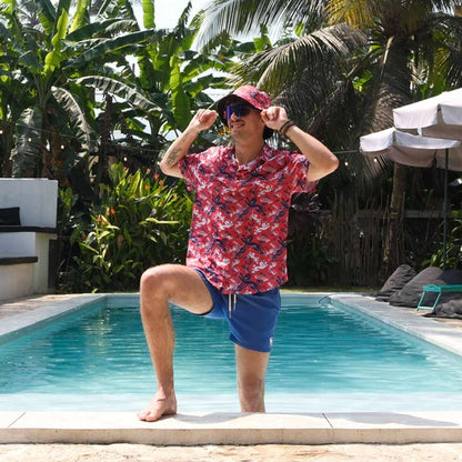 Wearing the Red England FC Hawaiian Shirt, sunglasses, blue shorts, and a matching hat, a person poses playfully by a pool amidst lush tropical plants and palm trees.