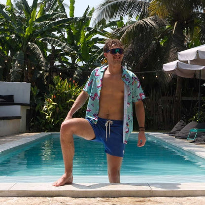 A man in the Flamingo Fandango Shirt, blue swim shorts, and sunglasses stands confidently at the pool’s edge, one foot on the stone border. Lush tropical plants and palm trees create a vibrant backdrop for his bold shirt style.