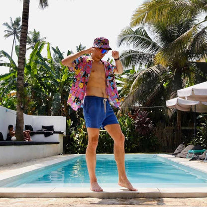 A man in a colorful shirt, blue shorts, and the Trippy Space Bucket Hat poses by a pool with tropical plants and palm trees. People relax under an umbrella in the background, showcasing this vibrant festival accessory.