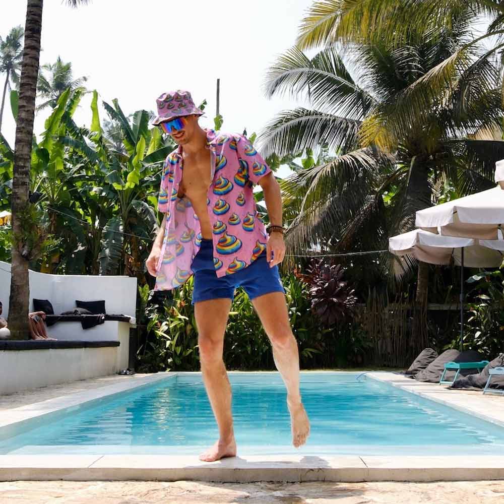 Wearing a Unicorn Poo Shirt, blue shorts, and a bucket hat, a man playfully steps beside a pool surrounded by tropical plants and palm trees on a sunny day.