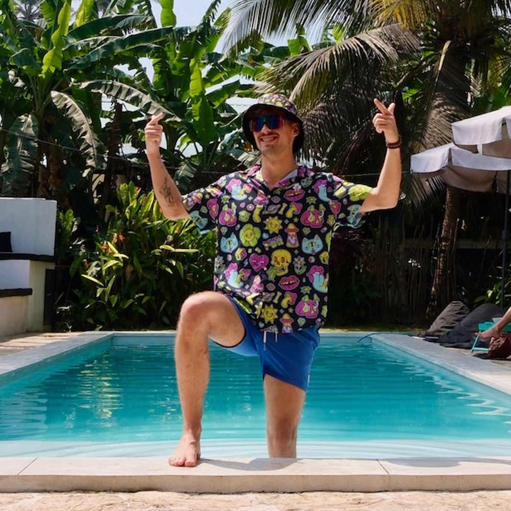 Wearing the Black Out Mushroom Bucket Hat, a man in a vibrant print shirt and blue shorts strikes a confident pose by the pool, raising one knee and pointing upward amid tropical plants and palm trees on a sunny day.