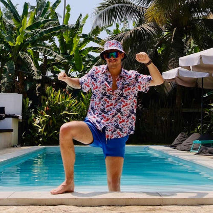 Wearing an England Football Bucket Hat, a man in a colorful shirt, blue shorts, and sunglasses playfully poses by a sunlit pool surrounded by tropical plants and palm trees.