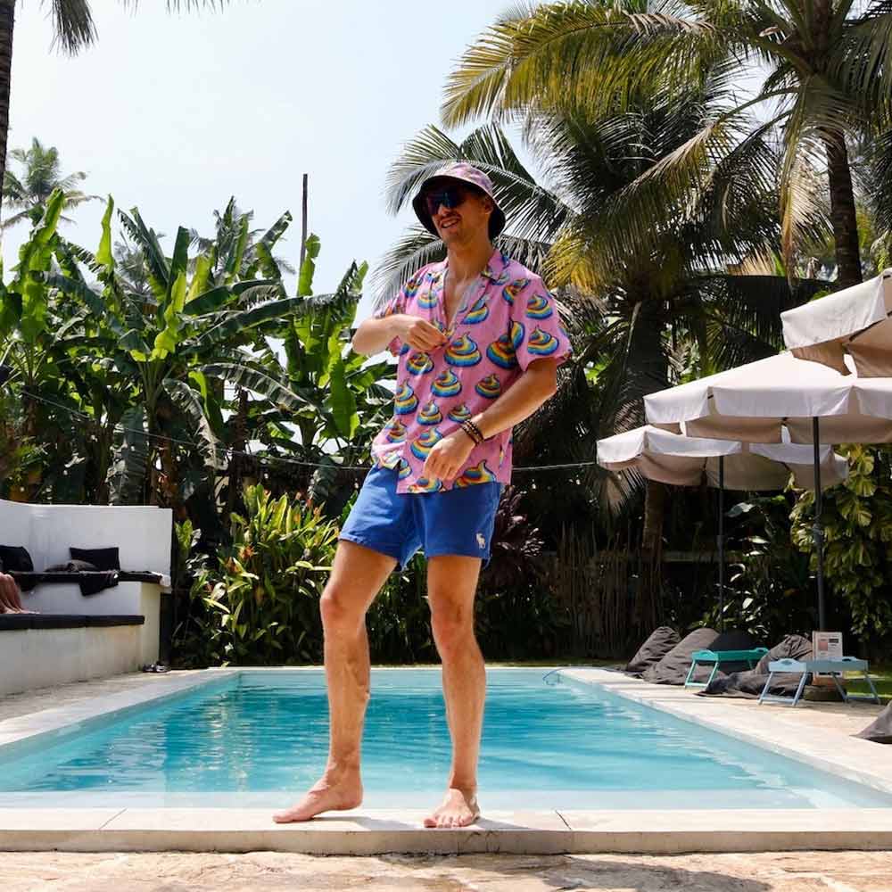 A man in a Unicorn Poo Shirt, blue shorts, and a bucket hat strikes a playful pose by the pool, with tropical plants and palm trees around him on a sunny day.