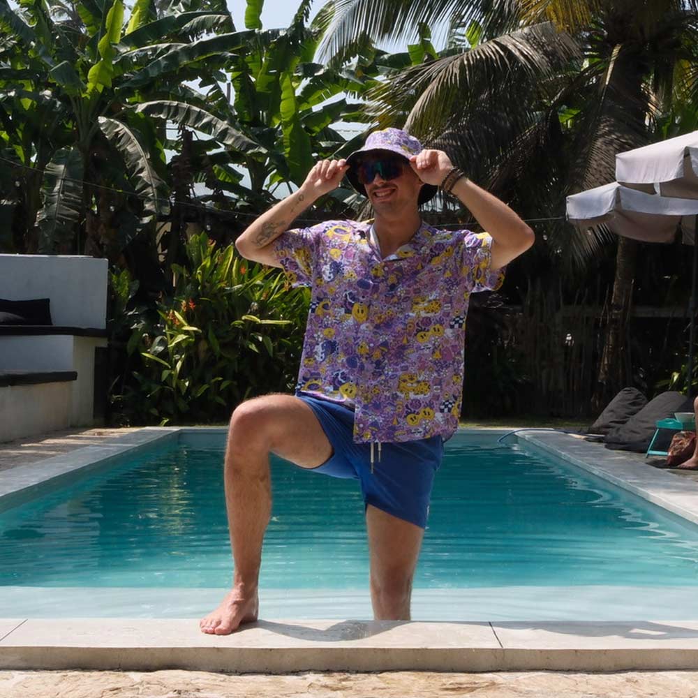 Wearing a Magical Psychedelic Bucket Hat, colorful shirt, and festival fashion blue shorts, a person poses poolside with one foot on the edge and smiles outdoors, surrounded by lush green plants and palm trees.