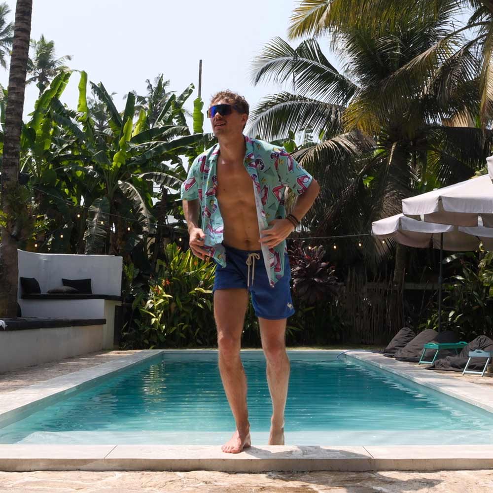 A man wearing the Flamingo Fandango Shirt, blue swim trunks, and sunglasses stands by a swimming pool amid lush tropical plants and palm trees. Lounge chairs and umbrellas can be seen in the background.