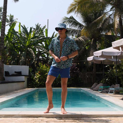 A man in blue swim shorts, a patterned shirt, and the Turquoise Tempest Leopard Print Bucket Hat stands by a swimming pool surrounded by tropical plants and palm trees on a sunny day.