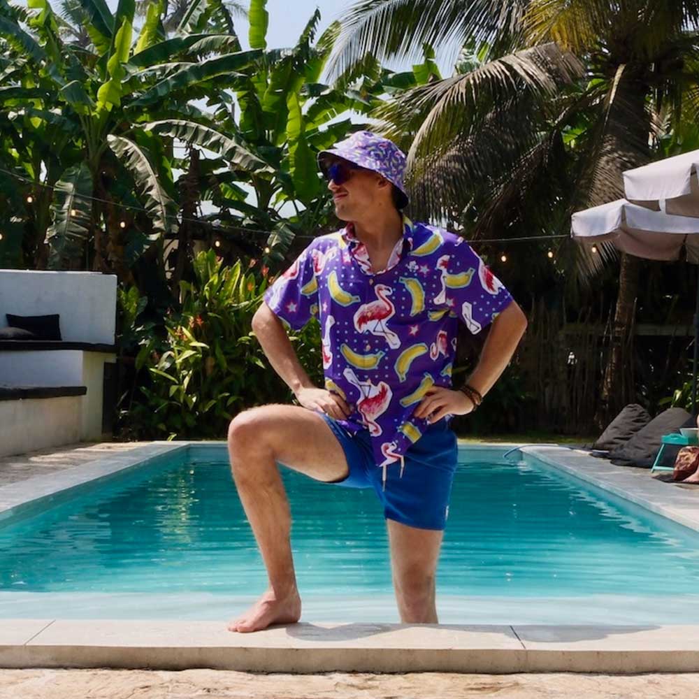 Wearing the Bananas For Flamingos Bucket Hat, a person in a colorful shirt and blue shorts poses with one foot on the pool edge, surrounded by tropical plants and palm trees in a sunny outdoor pool setting.