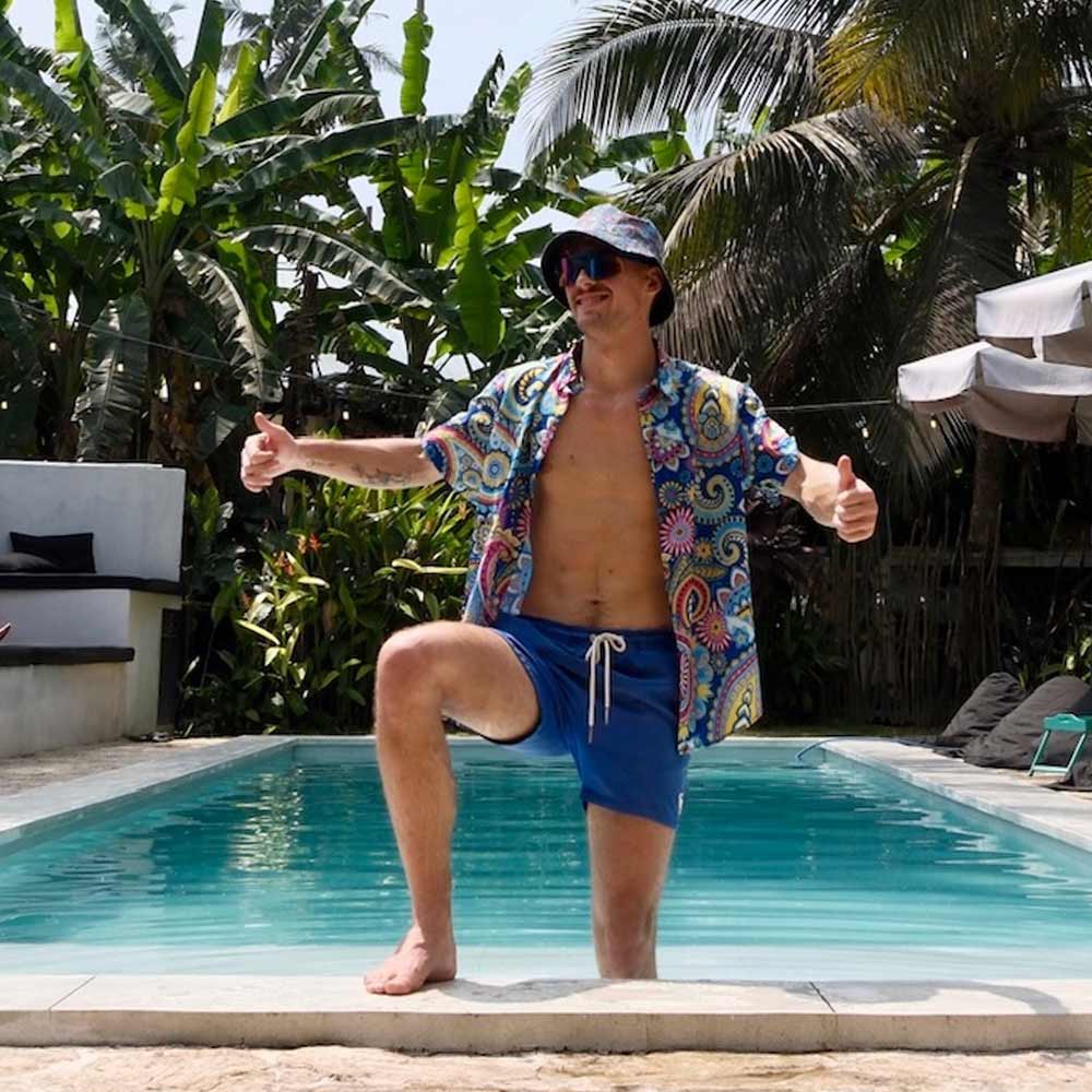 Wearing a Psychedelic Explorer Shirt, blue shorts, and a bucket hat, a man stands by the pool with one knee on the edge and gives two thumbs up. Palm trees and tropical plants surround the scene.