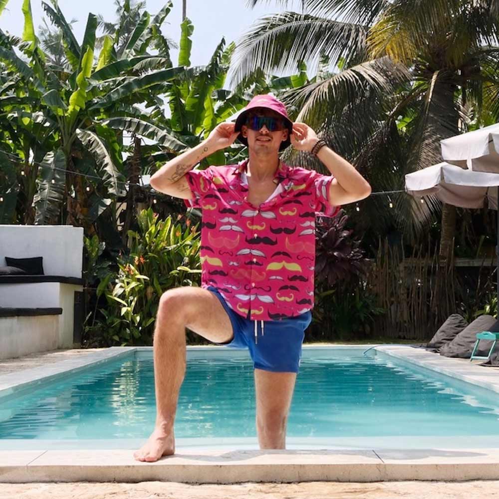 Wearing a Movember Bucket Hat, a man in a pink mustache-print shirt and blue shorts poses with one knee up at the edge of a pool, surrounded by tropical plants and palm trees.