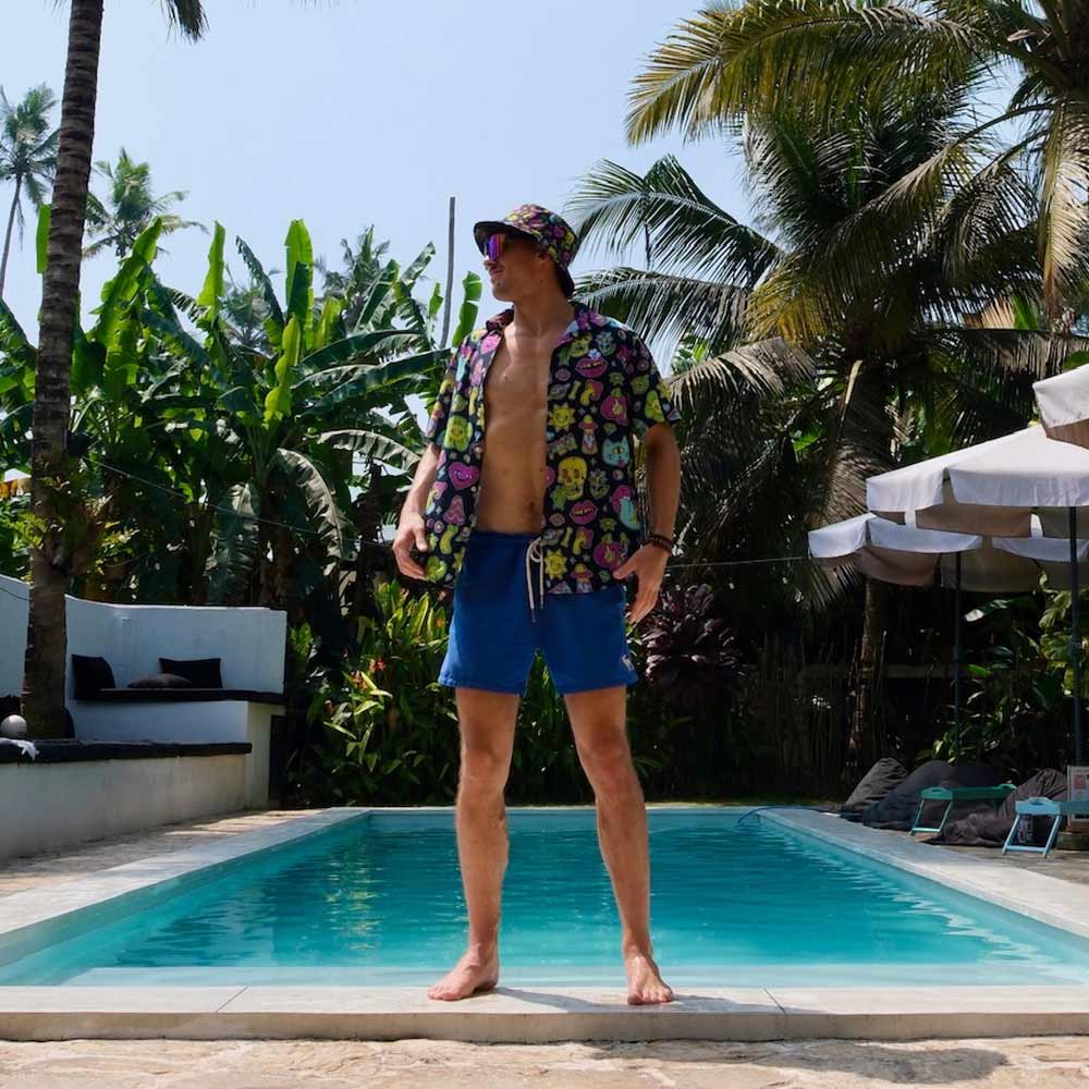 A man in a colorful shirt, blue shorts, and the Black Out Mushroom Bucket Hat stands barefoot by a pool, surrounded by tropical plants and palm trees under a sunny sky.