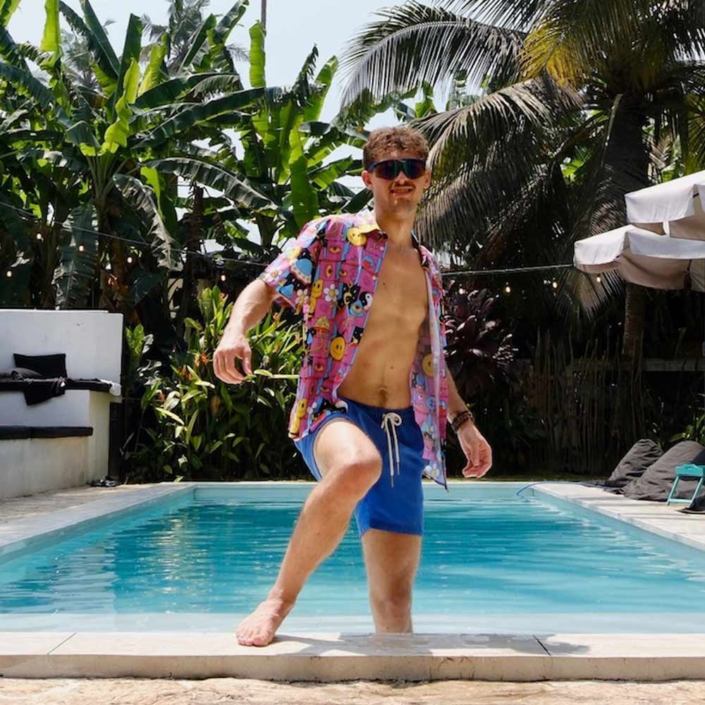 A man in sunglasses and blue swim trunks steps into a pool on a sunny day, surrounded by tropical plants, wearing the Trippy Space Shirt—a psychedelic magenta top that stands out amid lush palms.