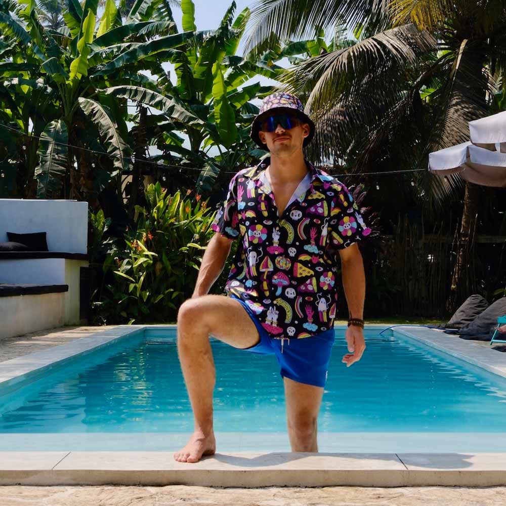 Wearing the Shroom Bucket Hat, sunglasses, a colorful shirt, and blue shorts, a man stands with one foot on the edge of a swimming pool surrounded by lush tropical plants and palm trees.