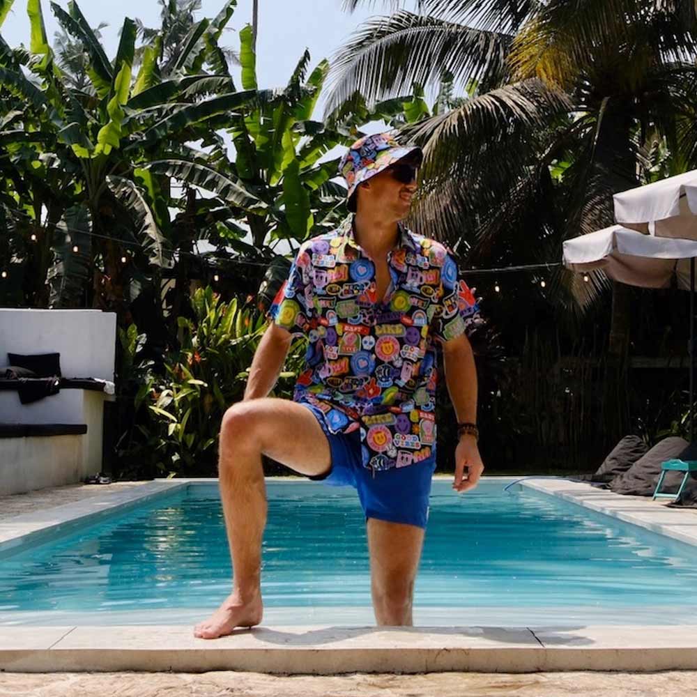 Wearing a Retro Sticker Bucket Hat, colorful patterned shirt, and blue shorts, a man stands with one foot in a swimming pool amid lush tropical plants and palm trees.