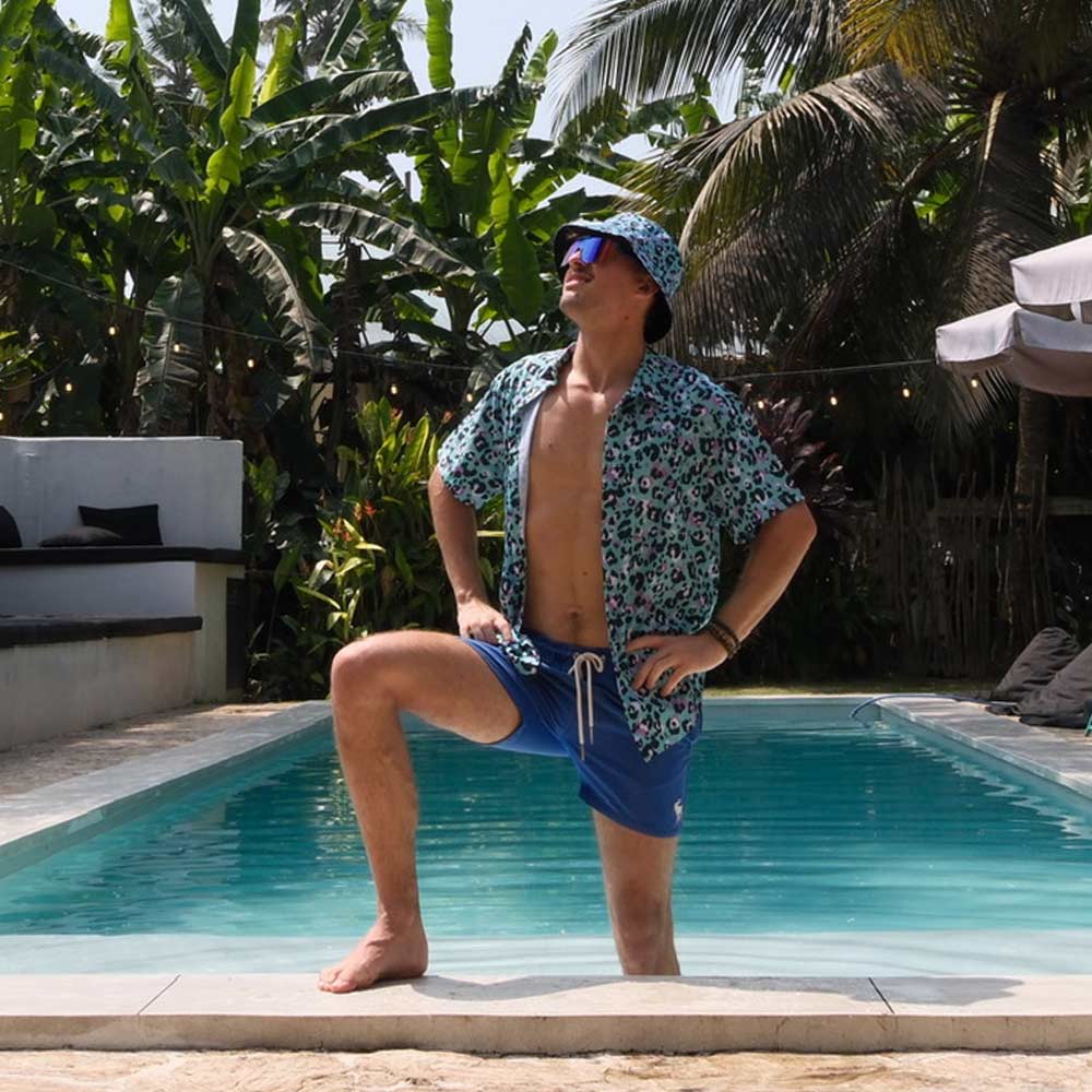 Wearing a Turquoise Tempest Leopard Print Bucket Hat, blue swim trunks, and a patterned shirt, a man stands confidently by the pool’s edge, surrounded by tropical plants and palm trees.