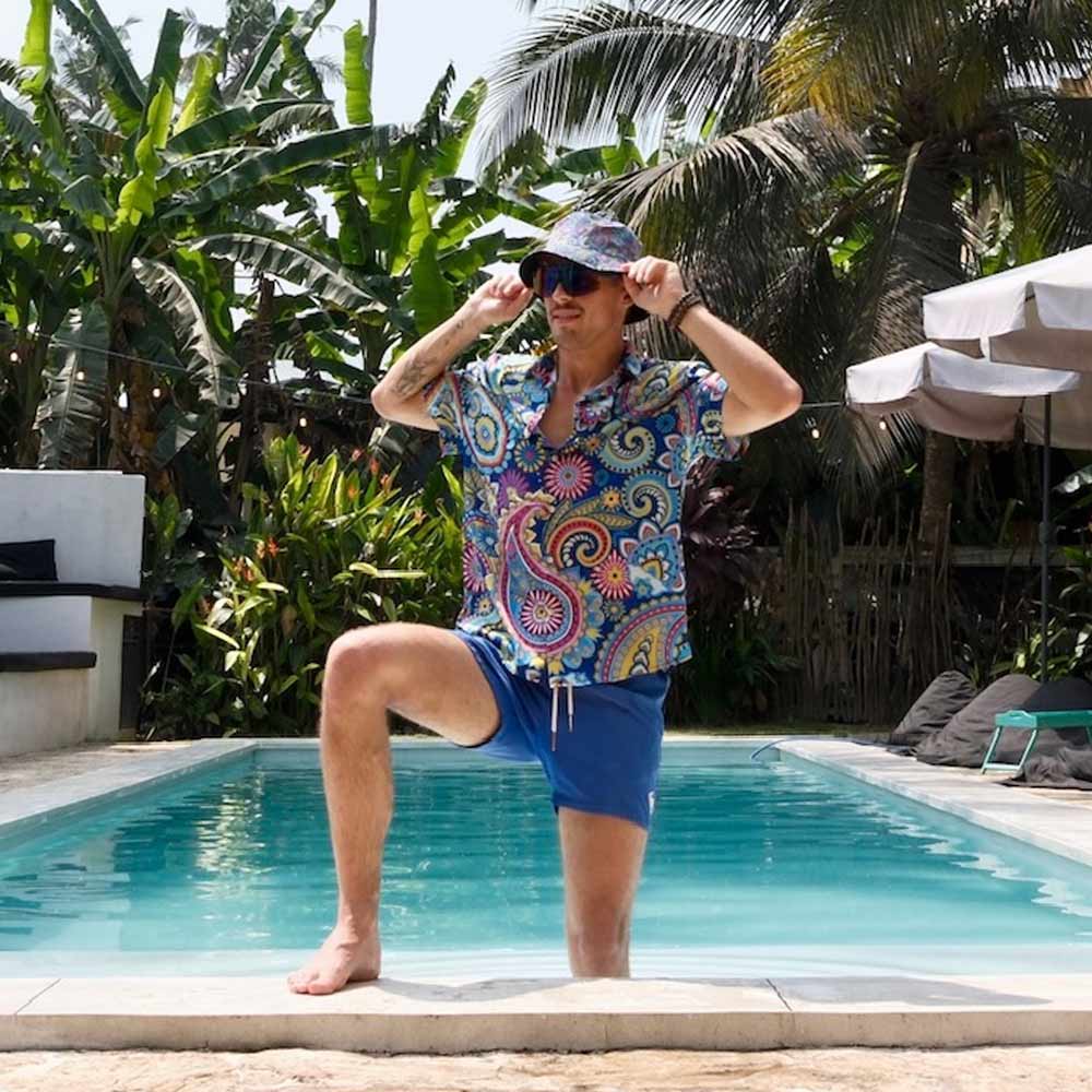 A man in a colorful Psychedelic Explorer Shirt, blue shorts, and a bucket hat poses playfully next to a pool, surrounded by vibrant tropical plants and palm trees.