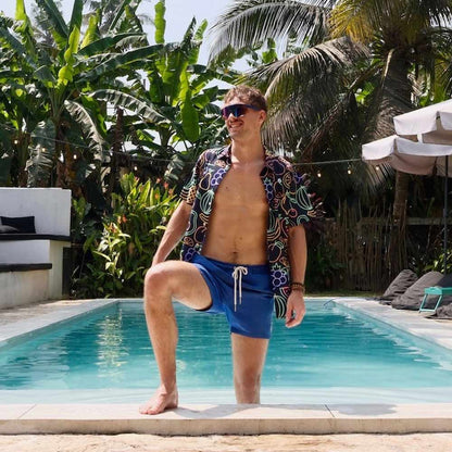 A man in sunglasses and a Fluorescent Fruit Fiesta Shirt with blue swim shorts kneels at the edge of a swimming pool, surrounded by palm trees and tropical plants.
