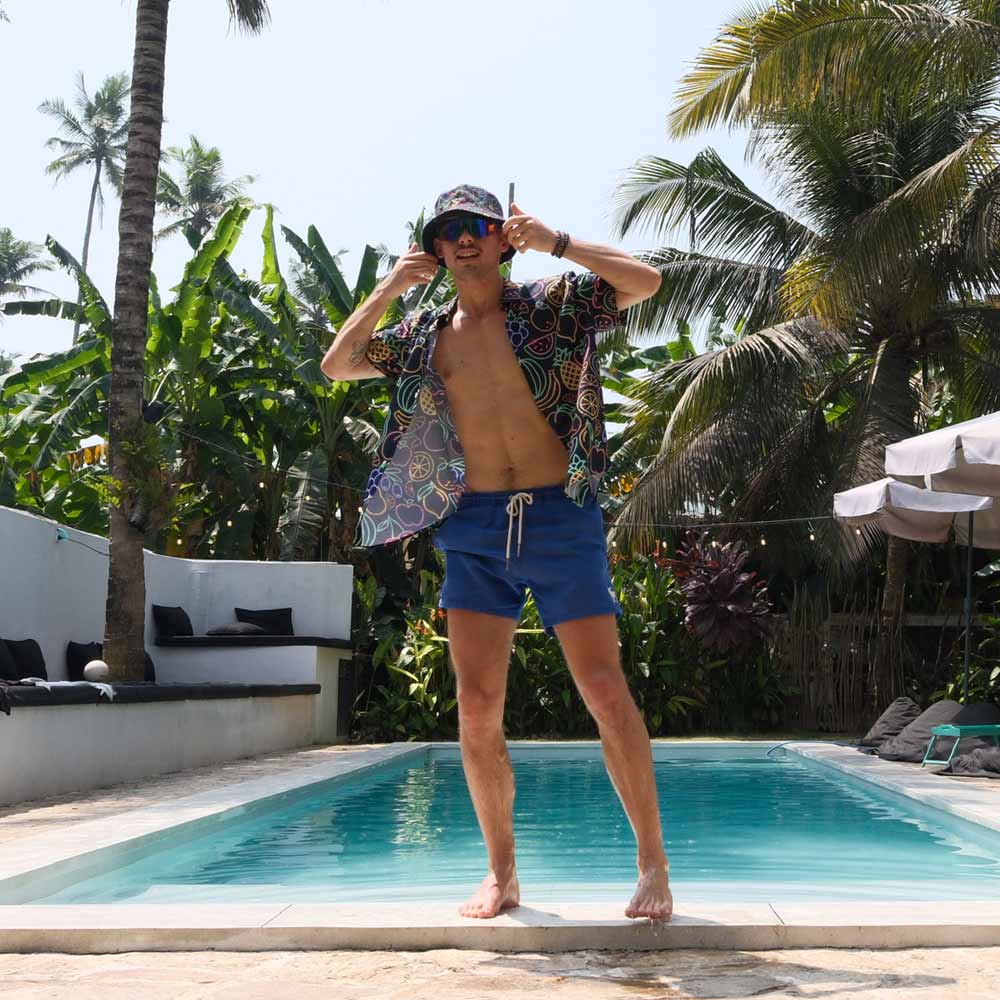 Wearing the Fluorescent Fruit Fiesta Bucket Hat, a man in blue swim shorts and an open patterned shirt poses playfully by a pool, surrounded by lush palm trees and greenery.