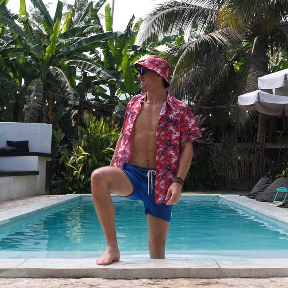 Wearing a red patterned shirt, blue swim shorts, and a Red England FC Away Bucket Hat, a person stands with one foot on the pools edge, surrounded by lush tropical plants and palm trees.