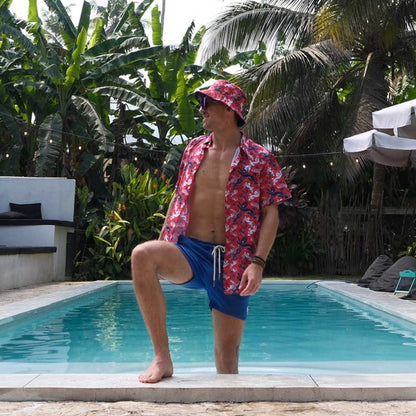 Wearing a red patterned shirt, blue swim shorts, and a Red England FC Away Bucket Hat, a person stands with one foot on the pools edge, surrounded by lush tropical plants and palm trees.