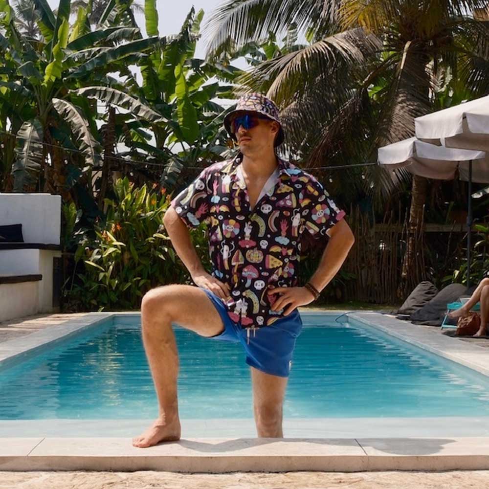 Wearing a Shroom Bucket Hat, blue shorts, and a vibrant patterned shirt, a man stands confidently with one leg on the pool edge and the other in the water, surrounded by lush tropical plants and palm trees.