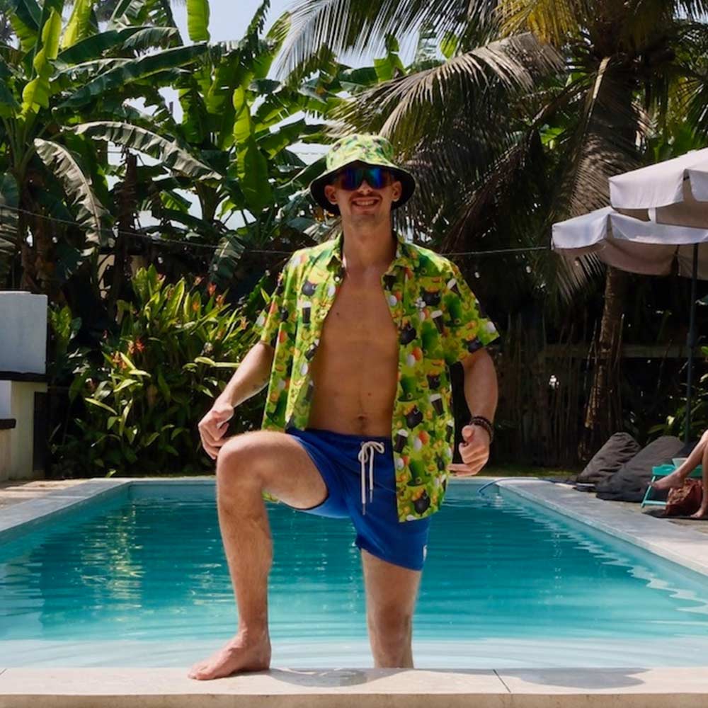 Wearing a colorful tropical shirt, blue shorts, and the St Patrick’s Day Bucket Hat, a person stands with one foot in a pool, surrounded by lush palm trees and greenery on a sunny day.