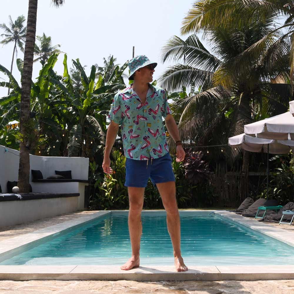 Wearing a Flamingo Fandango Bucket Hat, patterned shirt, and blue shorts, a man stands barefoot by a swimming pool, tropical plants and palm trees all around.