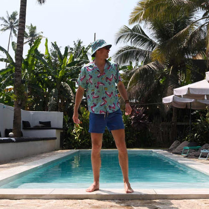 Wearing a Flamingo Fandango Bucket Hat, patterned shirt, and blue shorts, a man stands barefoot by a swimming pool, tropical plants and palm trees all around.