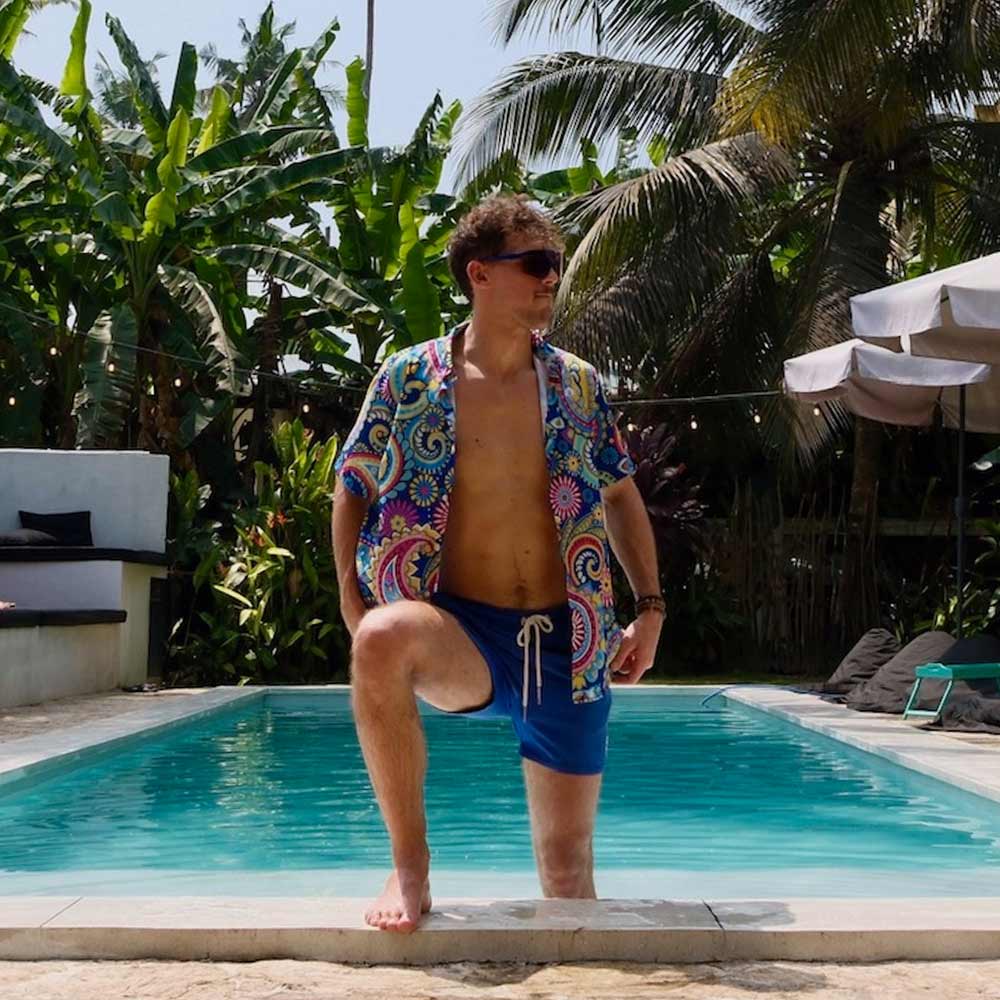 A man in the Psychedelic Explorer Shirt and sunglasses stands confidently with one leg on a backyard pools edge, surrounded by lush tropical plants and palm trees.
