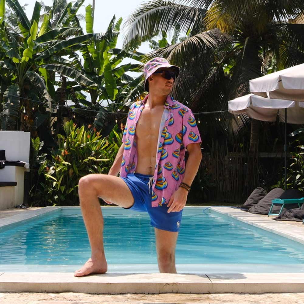 Wearing blue swim trunks, a Unicorn Poo Shirt with pink patterns, and a matching bucket hat, a man poses confidently with one foot on the pool’s edge, surrounded by tropical plants and palm trees.