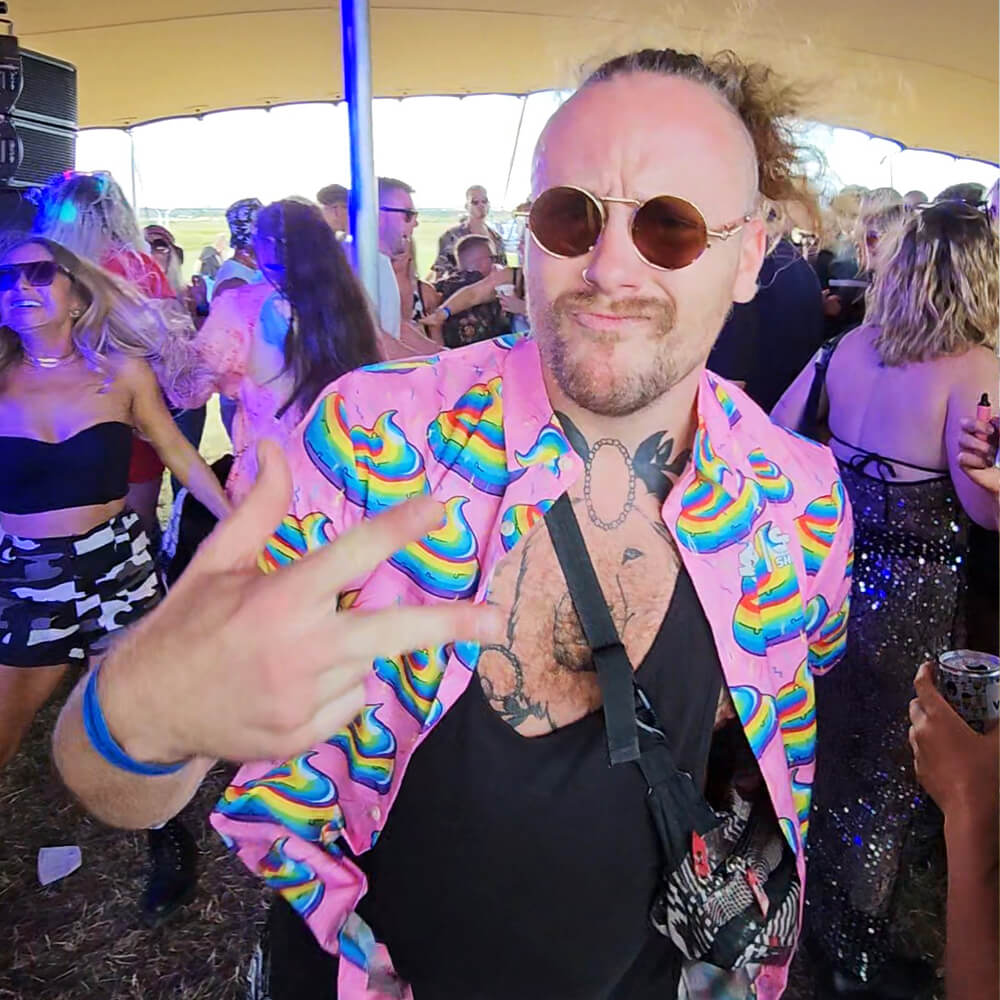 A man wearing round sunglasses and a colorful shirt with rainbow patterns makes a peace sign at an outdoor party, surrounded by dancing people under a canopy.