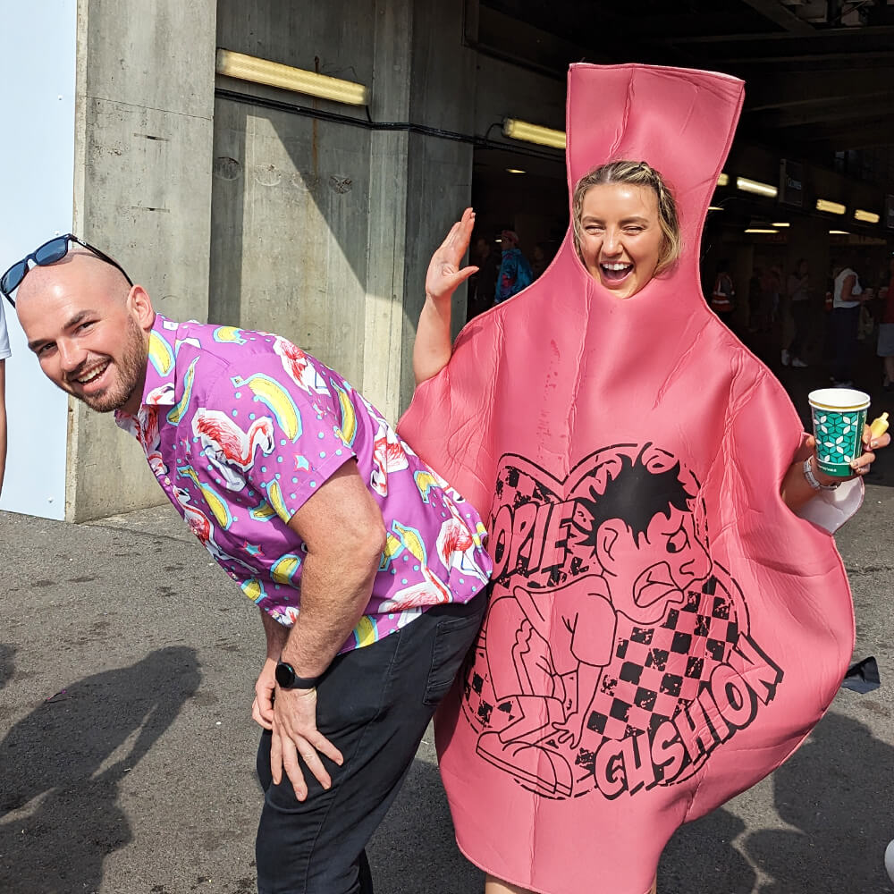A man in a colorful flamingo shirt laughs as a woman dressed in a large whoopee cushion costume pretends to press on his backside; both are having fun at an outdoor event.