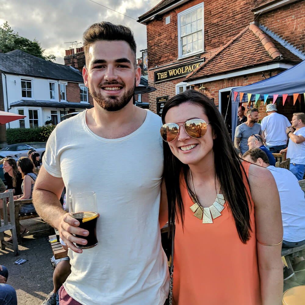 A smiling man with a beard holds a drink and stands next to a smiling woman in sunglasses and an orange top. They are outdoors at a lively pub garden with people and colorful bunting in the background.