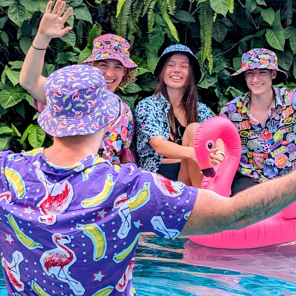 Four people in colorful, patterned shirts and bucket hats laugh and pose by a pool, with one sitting on an inflatable pink flamingo and lush green leaves in the background.