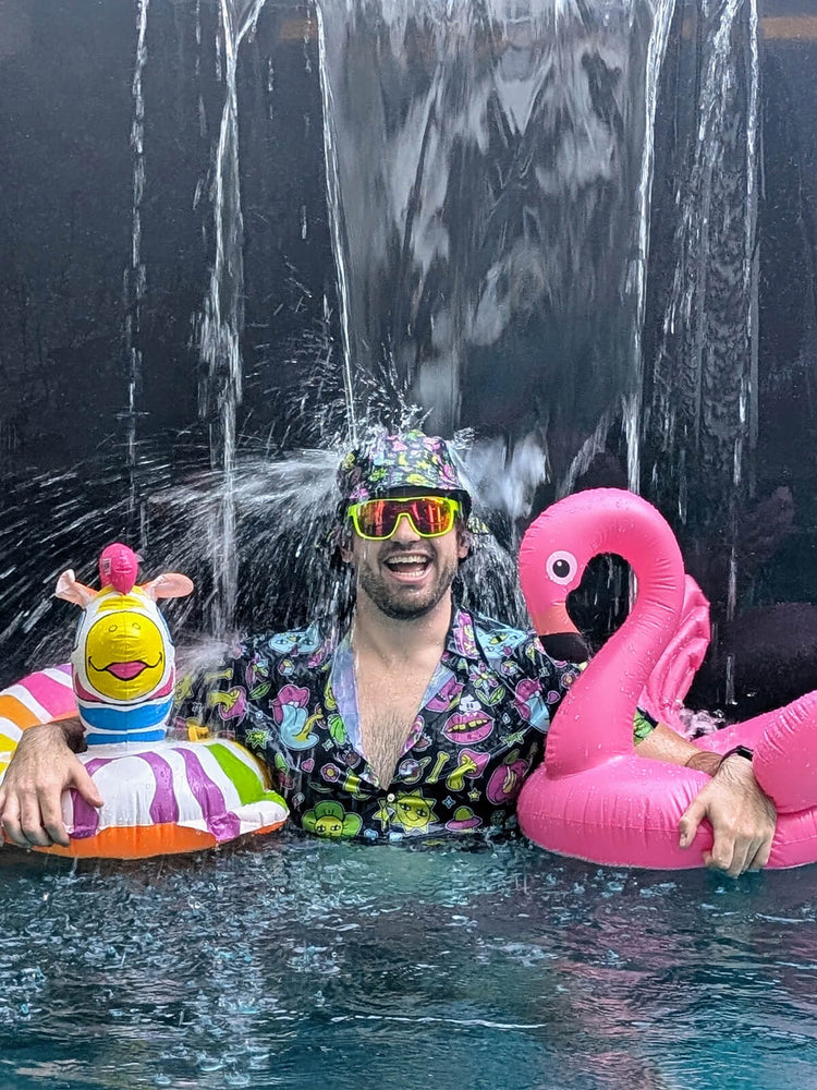 A man in colorful clothes and sunglasses stands in a pool under a waterfall, holding a striped float with a duck toy and a pink flamingo float, smiling happily.